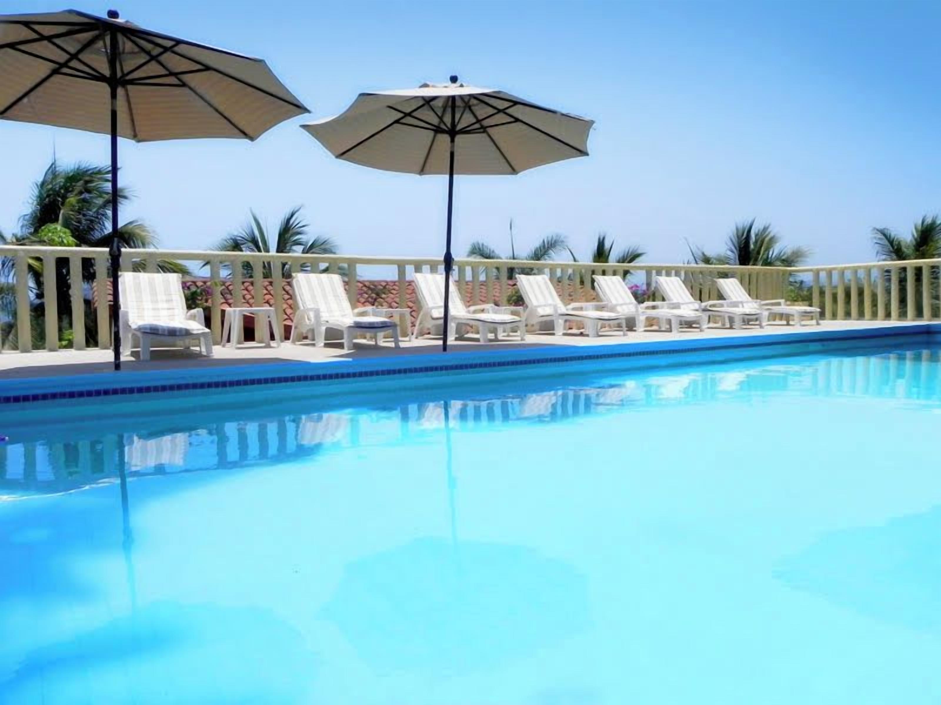 Piscina con sillas de descanso y sombrillas, rodeada por una valla y palmeras.