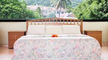 A bedroom with a bed, two nightstands, and a view of a lush green landscape with trees and a building in the background.