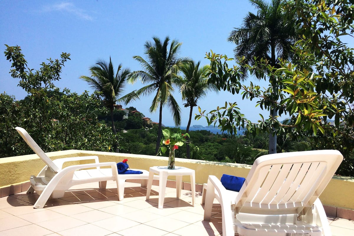 Een terras met witte loungestoelen en een kleine tafel, met uitzicht op een weelderig landschap met palmbomen en zee.