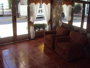 Habitación con pared de piedra, piso de madera y vista a la calle a través de amplios ventanales con cortinas.