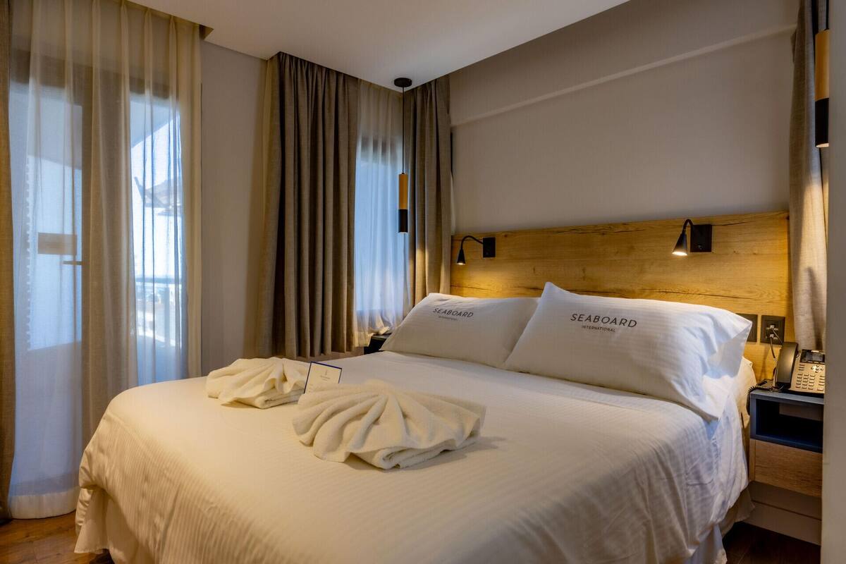 Camera d'albergo con un letto grande, due cuscini con la scritta 'SEABOARD INTERNATIONAL', una testiera in legno, un comodino con una lampada e una finestra con tende.