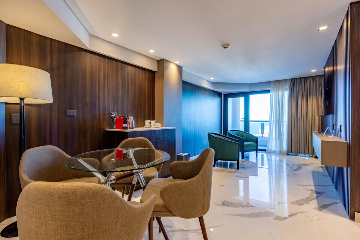 Una camera d'albergo moderna con un tavolo da pranzo in vetro, sedie confortevoli, un angolo caffè e vista sul mare attraverso ampie finestre.