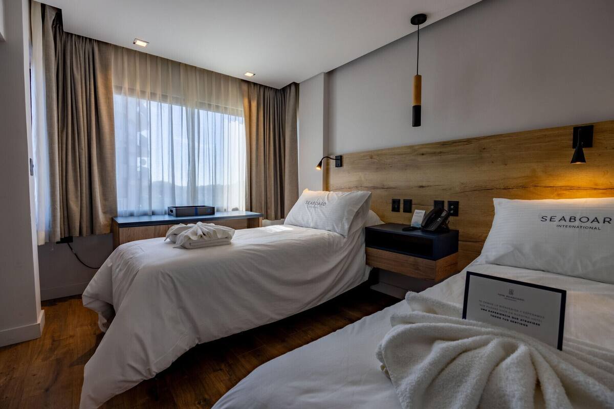 Camera d'albergo con due letti, una testiera in legno, una scrivania con un computer e una finestra con tende leggere.