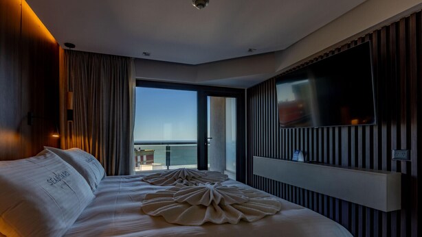 Ein Hotelzimmer mit einem großen Bett, einem flachbildfernseher an der Wand und Meerblick durch ein großes Fenster.