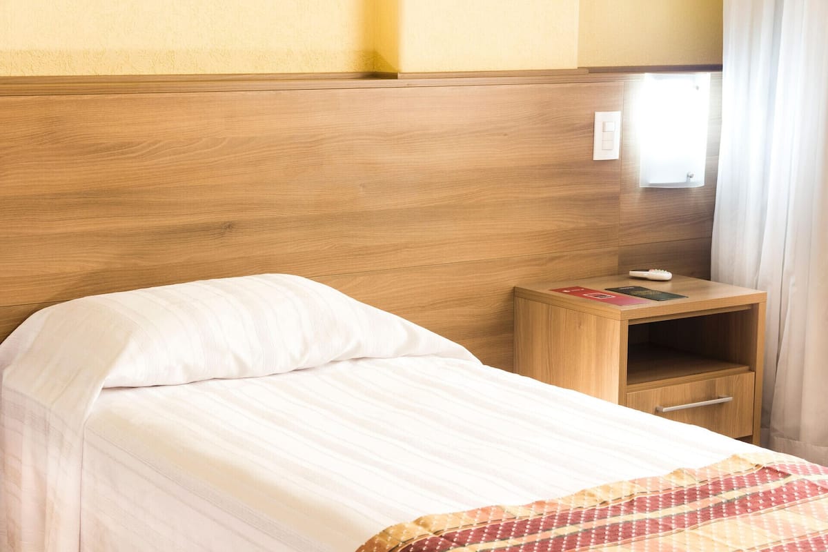 A hotel room with a wooden headboard, a single bed with white bedding, a small wooden nightstand with a remote control and a book, and a window with curtains.