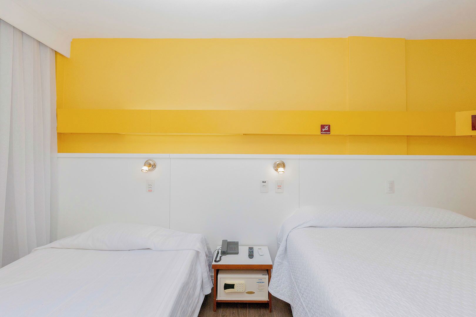 Une chambre d’hôtel avec deux lits simples, une table de chevet avec un téléphone et une radio, et un mur d’accent jaune.