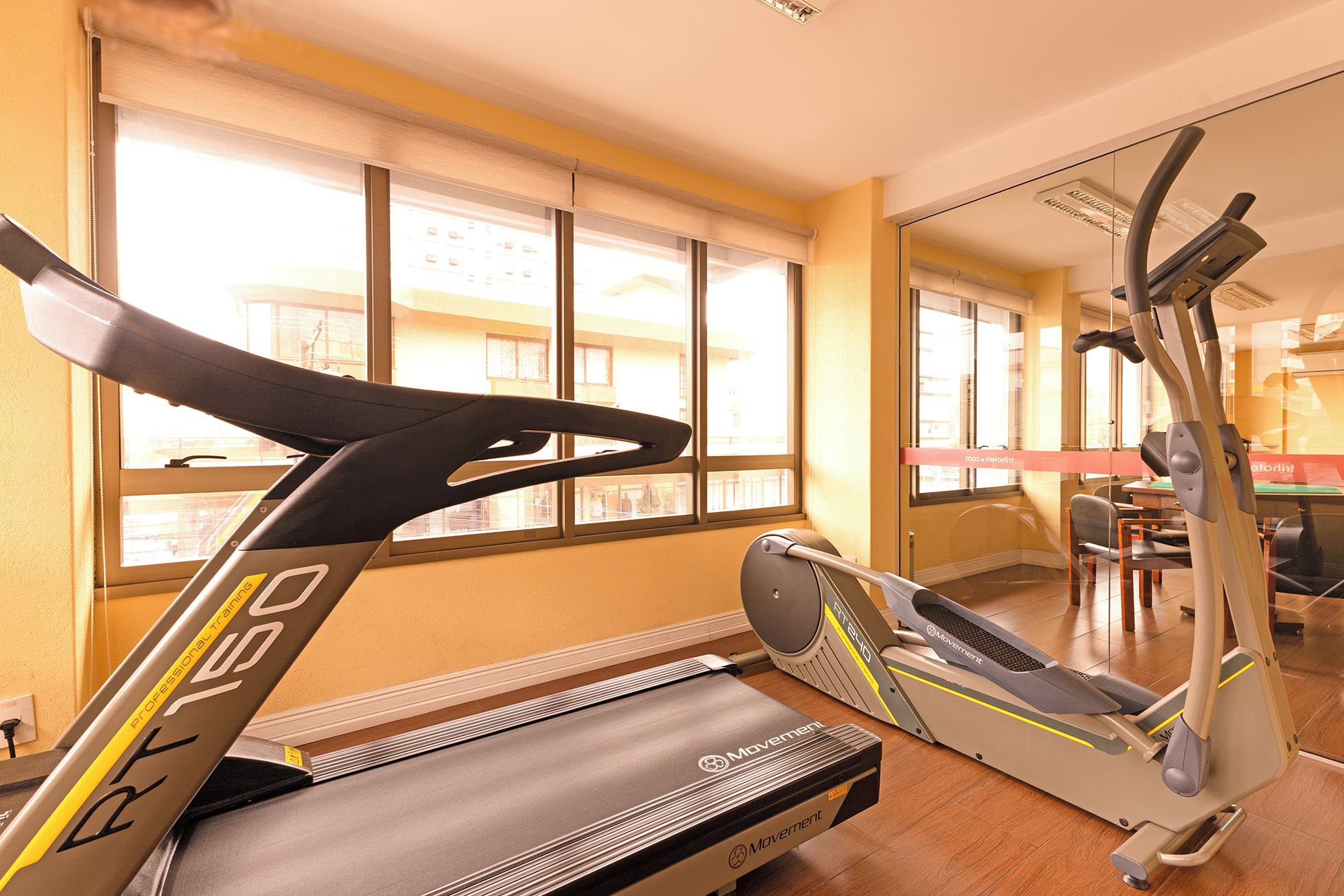 Un gimnasio con cinta de correr y máquina elíptica, ventanas amplias y un espejo que refleja la sala.