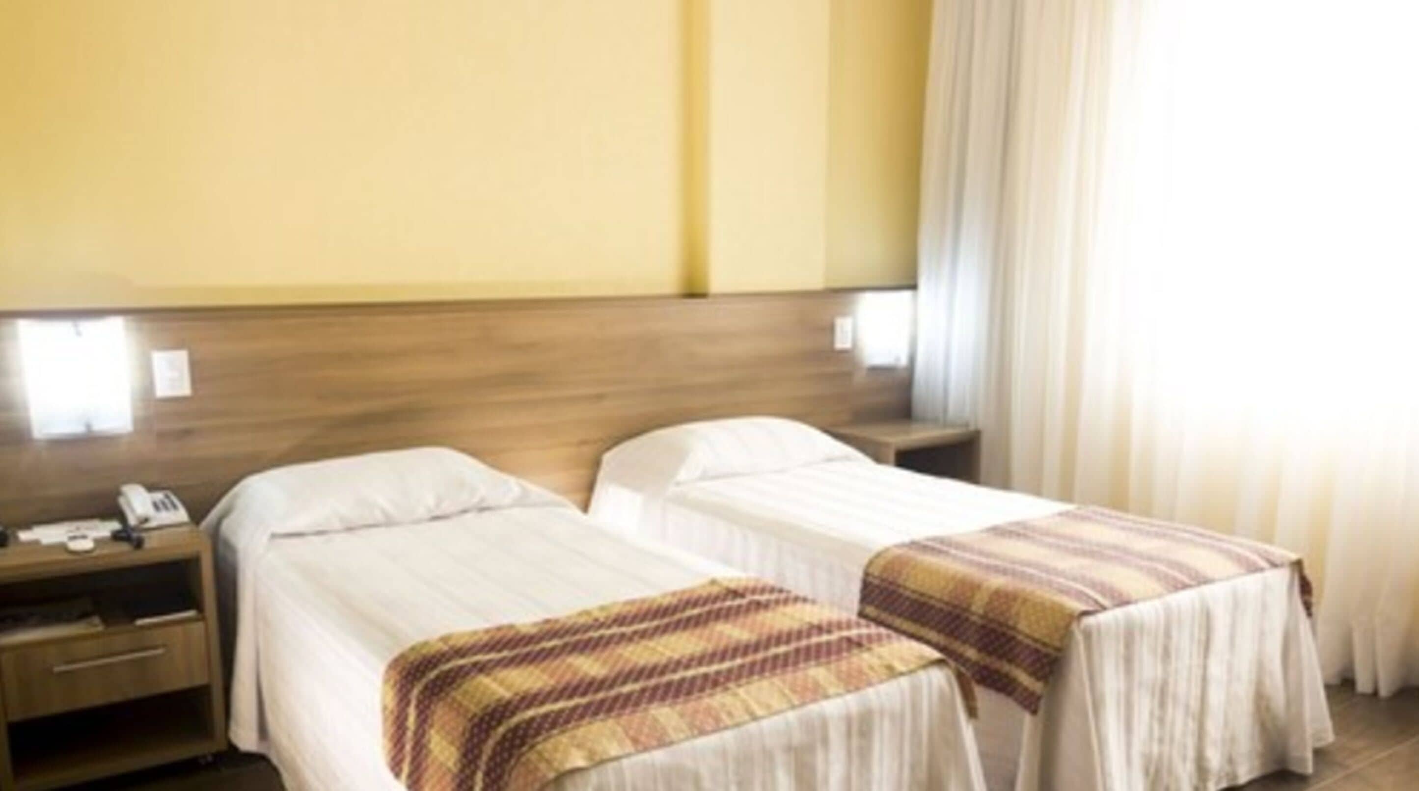 Habitación de hotel con dos camas, cabeceros de madera, mesitas de noche con lámparas y una ventana con cortinas.