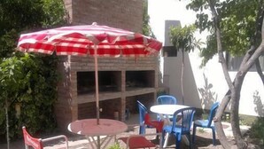 An outdoor patio with a round table and blue chairs, a red and white checkered umbrella, and a brick wall with greenery.