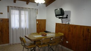 A dining area with a wooden table and chairs, a television mounted on the wall, and a window with curtains.
