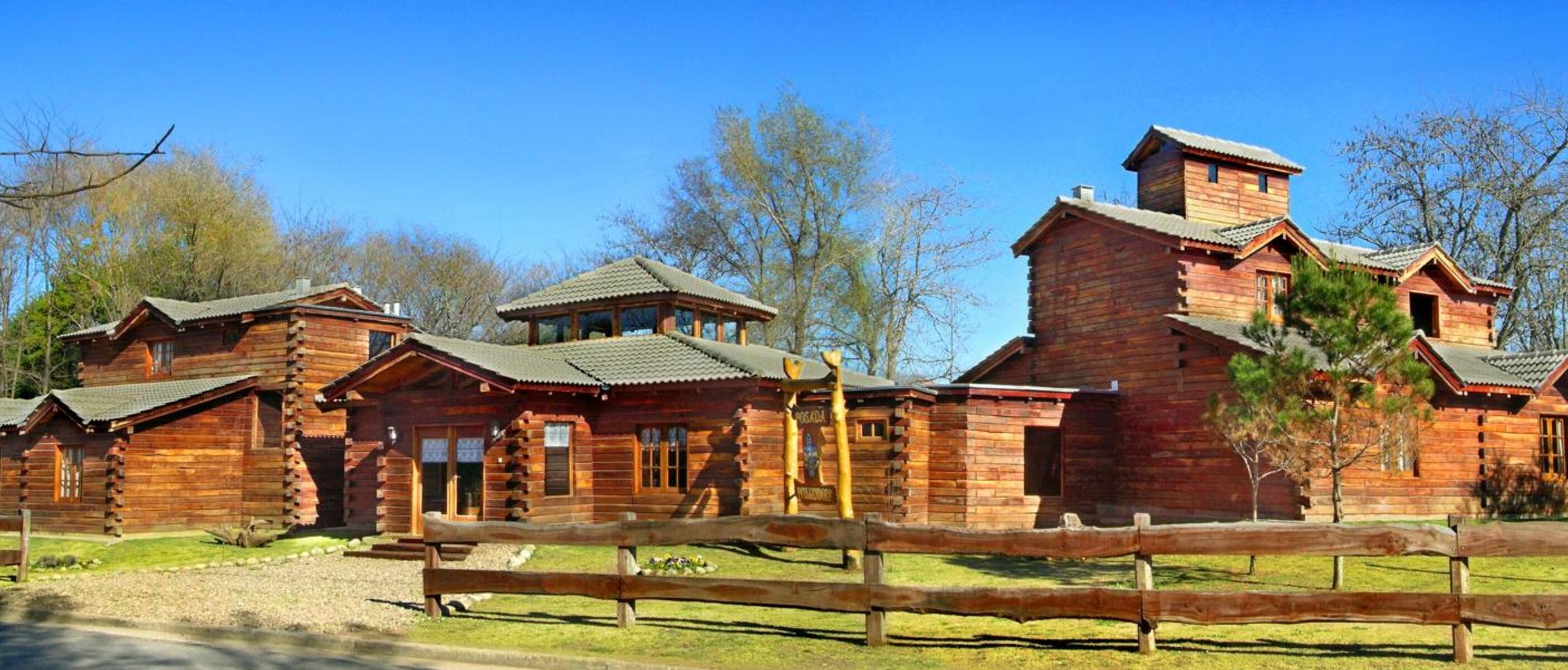 A row of wooden log cabins with a rustic design, featuring multiple chimneys and a wooden fence in the foreground.