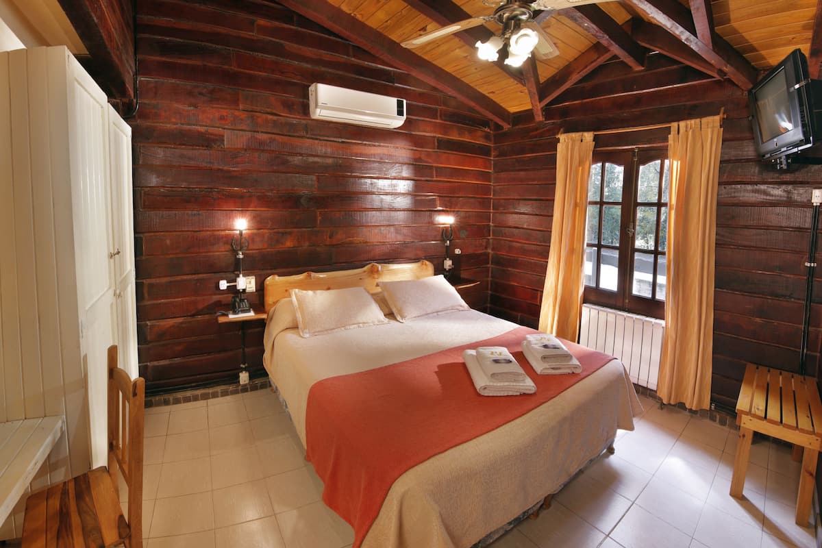 Un dormitorio de cabaña de madera con una cama, lámparas de noche, un ventilador de techo y un televisor montado en la pared.