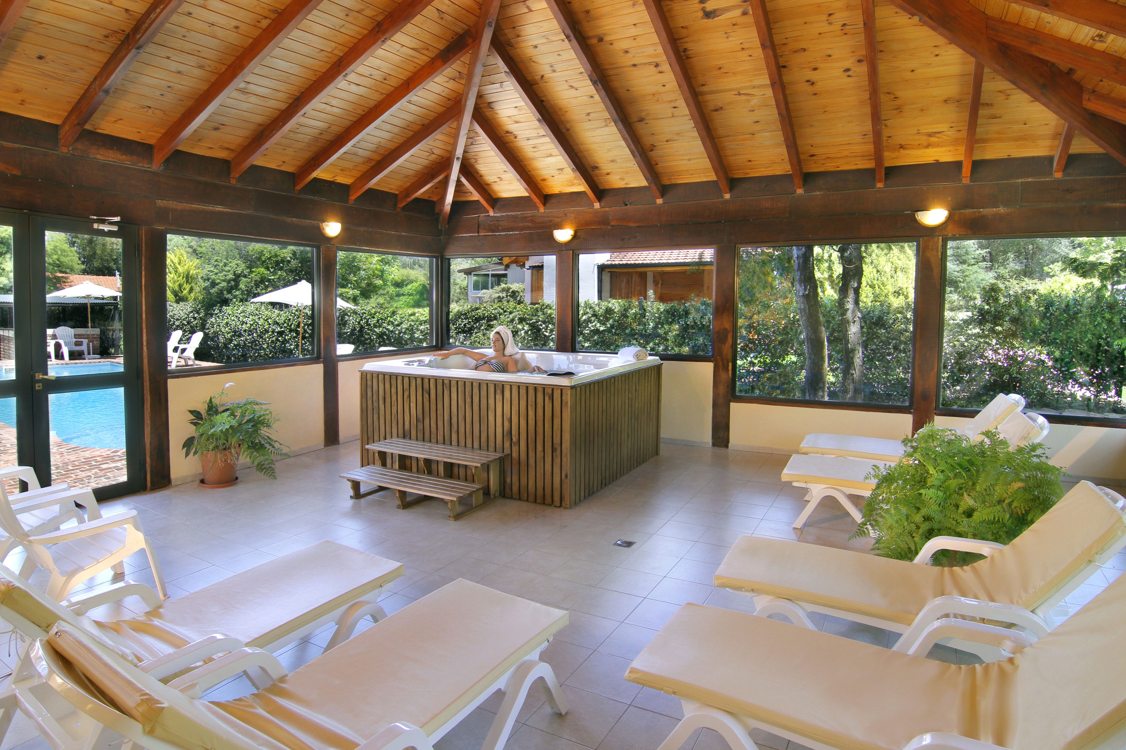 A covered outdoor area with wooden beams, a hot tub, lounge chairs, and a view of the pool and greenery.