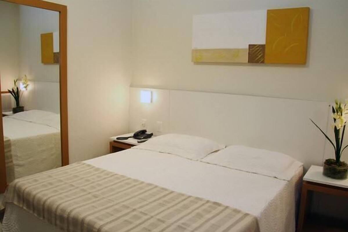 A hotel room with a bed, a bedside table with a lamp, a mirror, and a wall-mounted artwork.