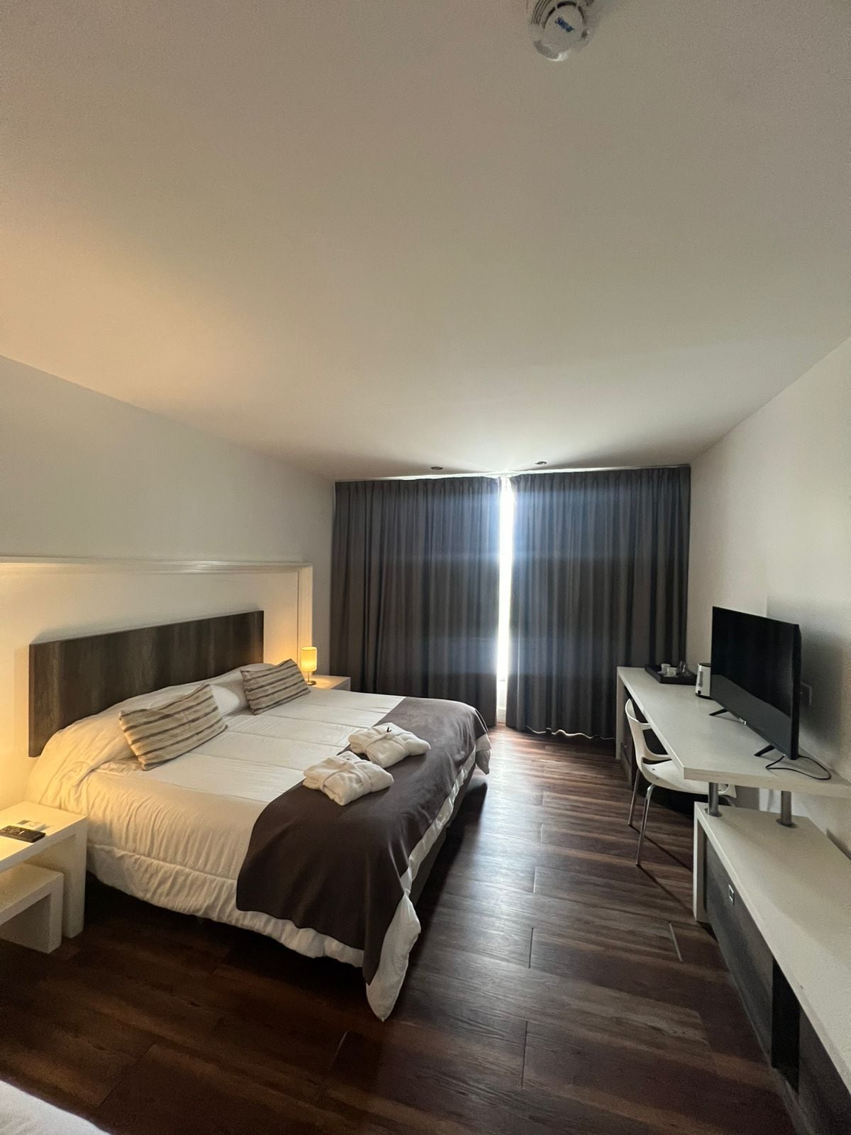 A hotel room with a bed, bedside table, TV, and a desk with a chair.