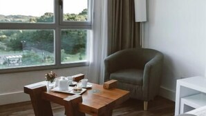 A hotel room with a large window, a wooden table with a vase and teapot, a green armchair, and a white bedside table. - Apart & Hotel Posta Natural (Tandil)