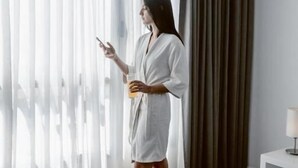 A person in a white robe standing by a window with sheer curtains, holding a smartphone and a glass of orange juice. - Apart & Hotel Posta Natural (Tandil)