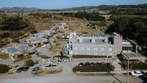An aerial view of a modern building complex with multiple parking areas, surrounded by hilly terrain and sparse vegetation. - Apart & Hotel Posta Natural (Tandil)