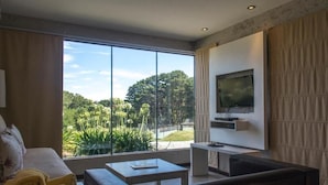 Uma sala de estar moderna com uma janela grande, uma televisão de tela plana na parede, um sofá, uma mesa de centro e uma cama bem arrumada com toalhas dobradas.