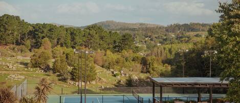 A tennis court with a covered seating area and a view of a hilly landscape.