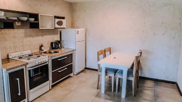 Cocina compacta con estufa a gas, horno, microondas y refrigerador. Hay una mesa de comedor con sillas y un pequeño estante sobre la estufa.