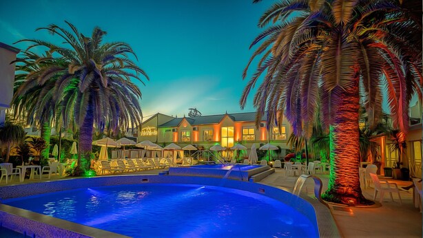 A pool area with lounge chairs and umbrellas, a building with a sign, and palm trees. - Alto Verde Suites and Apart (Termas de Rio Hondo)