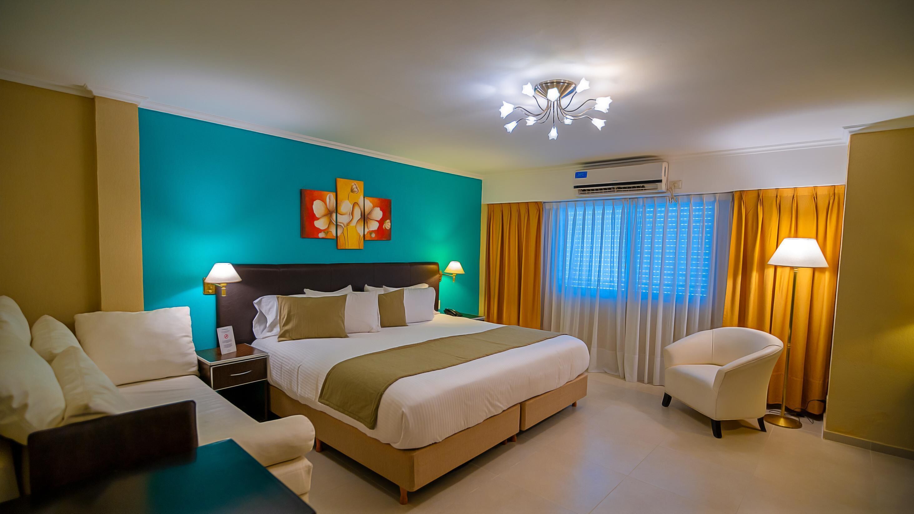 Habitación de hotel con cama, dos mesitas de noche, un escritorio, una silla, una lámpara, una obra de arte en la pared, un equipo de aire acondicionado y una ventana con cortinas.