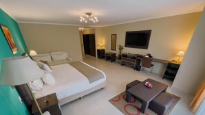 A hotel room with a bed, bedside table, lamp, desk, chair, TV, and a small seating area. - Alto Verde Suites and Apart (Termas de Rio Hondo)