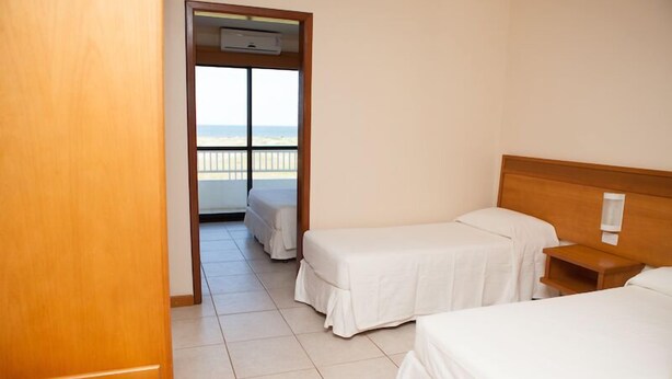 A hotel room with two single beds, a wooden headboard, a small bedside table, and a balcony with a view of the beach. - Nelson Praia Hotel (Rio Grande)