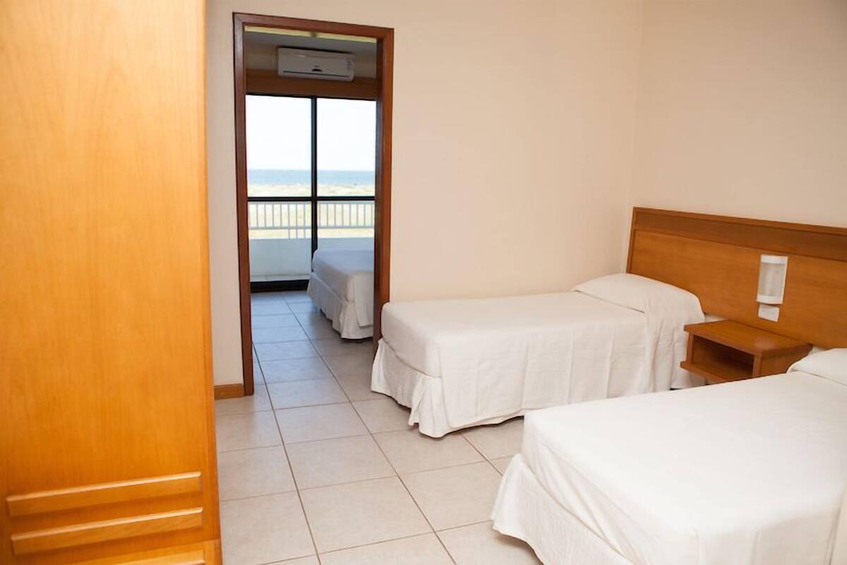 A hotel room with two single beds, a wooden headboard, a small bedside table, and a balcony with a view of the beach.