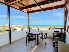 Ein überdachter Balkon mit Tisch und Stühlen, mit Blick auf einen Strand und das Meer.