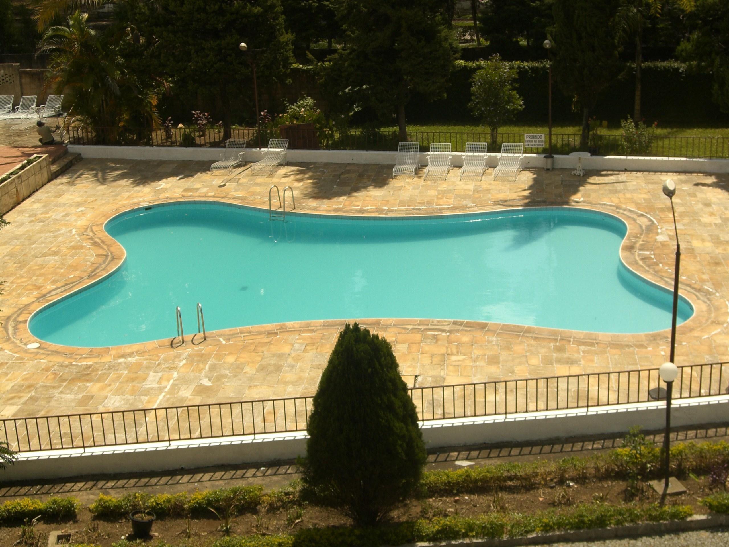 Piscina retangular com um deck ladrilhado, rodeada por uma cerca baixa e várias espreguiçadeiras.