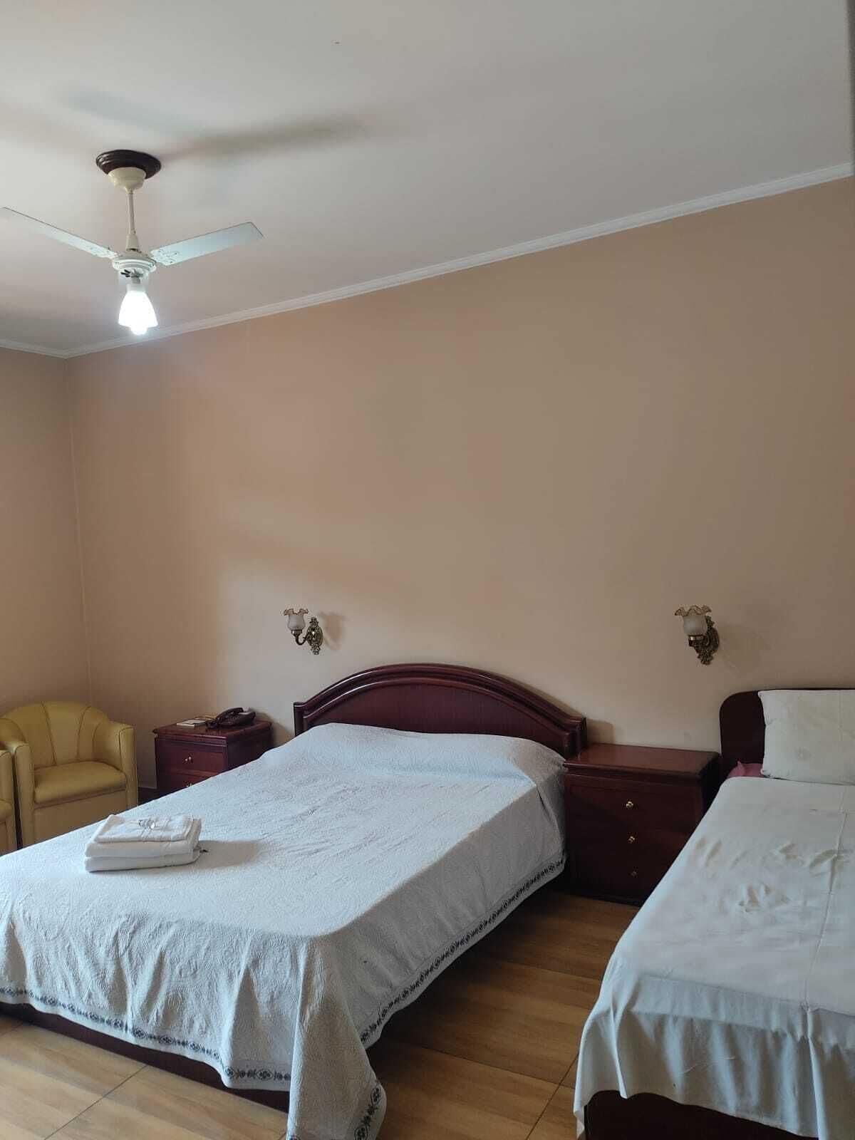 A hotel room with two beds, a ceiling fan, a bedside table with a lamp, and a chair.