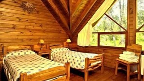 Uma cabana com duas camas de madeira, uma mesa pequena e uma janela com cortinas.