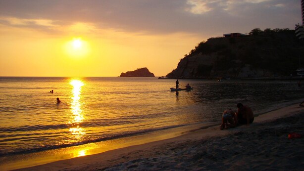 A beach scene at sunset with calm waters, a small boat, and people on the shore. - Apartamento Arena - Smr236a (Santa Marta)