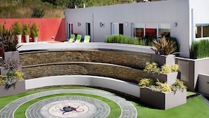 Un bâtiment moderne doté d’un mur extérieur incurvé, d’un patio circulaire orné d’une mosaïque et d’un aménagement paysager verdoyant.
