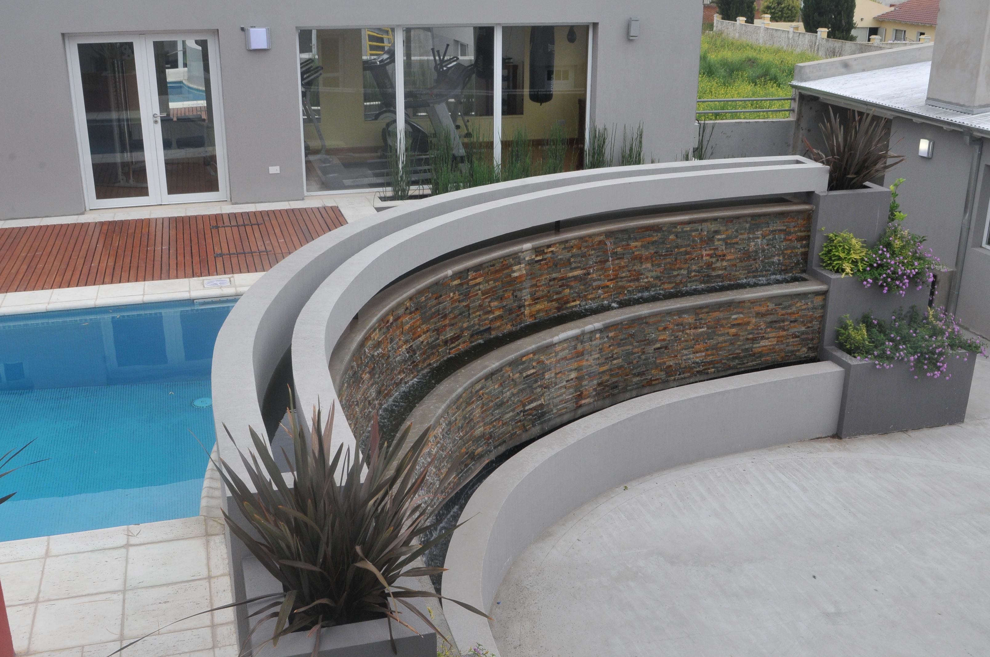 A modern pool area with a curved stone wall, a wooden deck, and potted plants.