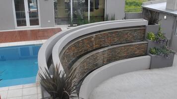 Un área de piscina moderna con un muro de piedra curvo, una terraza de madera y plantas en macetas.