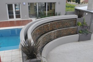 Un área de piscina moderna con un muro de piedra curvo, una terraza de madera y plantas en macetas.