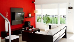 Un salon moderne avec un mur d’accent rouge, un téléviseur à écran plat, un canapé, une table basse et une grande fenêtre donnant sur un paysage verdoyant.