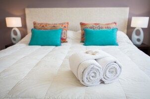 Una cama bien hecha con un edredón blanco, dos almohadas turquesa y una toalla blanca enrollada sobre la cama.