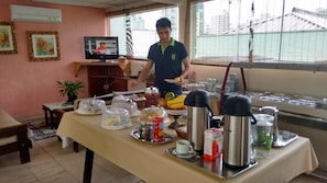 A breakfast buffet setup with a variety of food items and beverages, a television displaying a news program, and a window with a view of buildings and a bridge. - Hotel Podium (Sao Caetano do Sul)