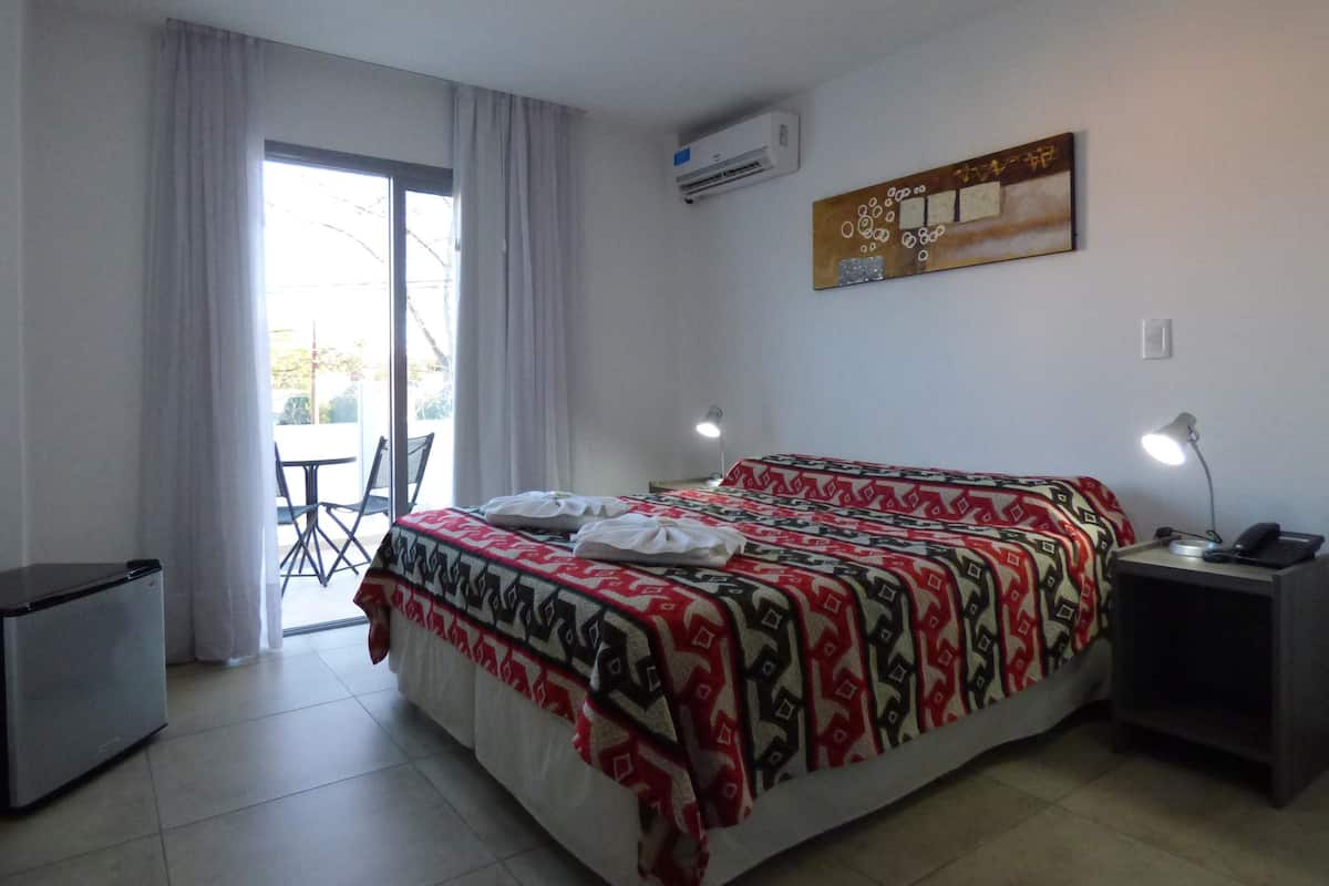 Una habitación de hotel con cama, escritorio, silla, aire acondicionado y ventana con cortinas.