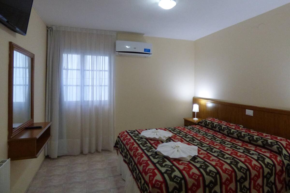Habitación de hotel con cama, mesita de noche, espejo, ventana con cortinas, equipo de aire acondicionado y luz de techo.