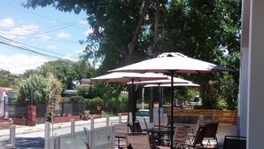An outdoor patio with tables and chairs, umbrellas, and a view of the street and trees. - Pinar del Lago (Villa Carlos Paz)