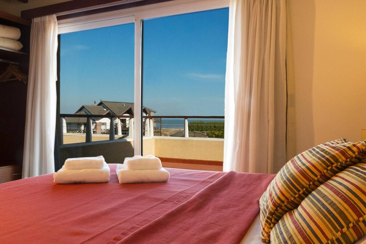 A hotel room with a bed, a red bedspread, two white towels, and a view of a building and landscape through large windows.