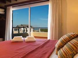 A hotel room with a bed, a red bedspread, two white towels, and a view of a building and landscape through large windows.