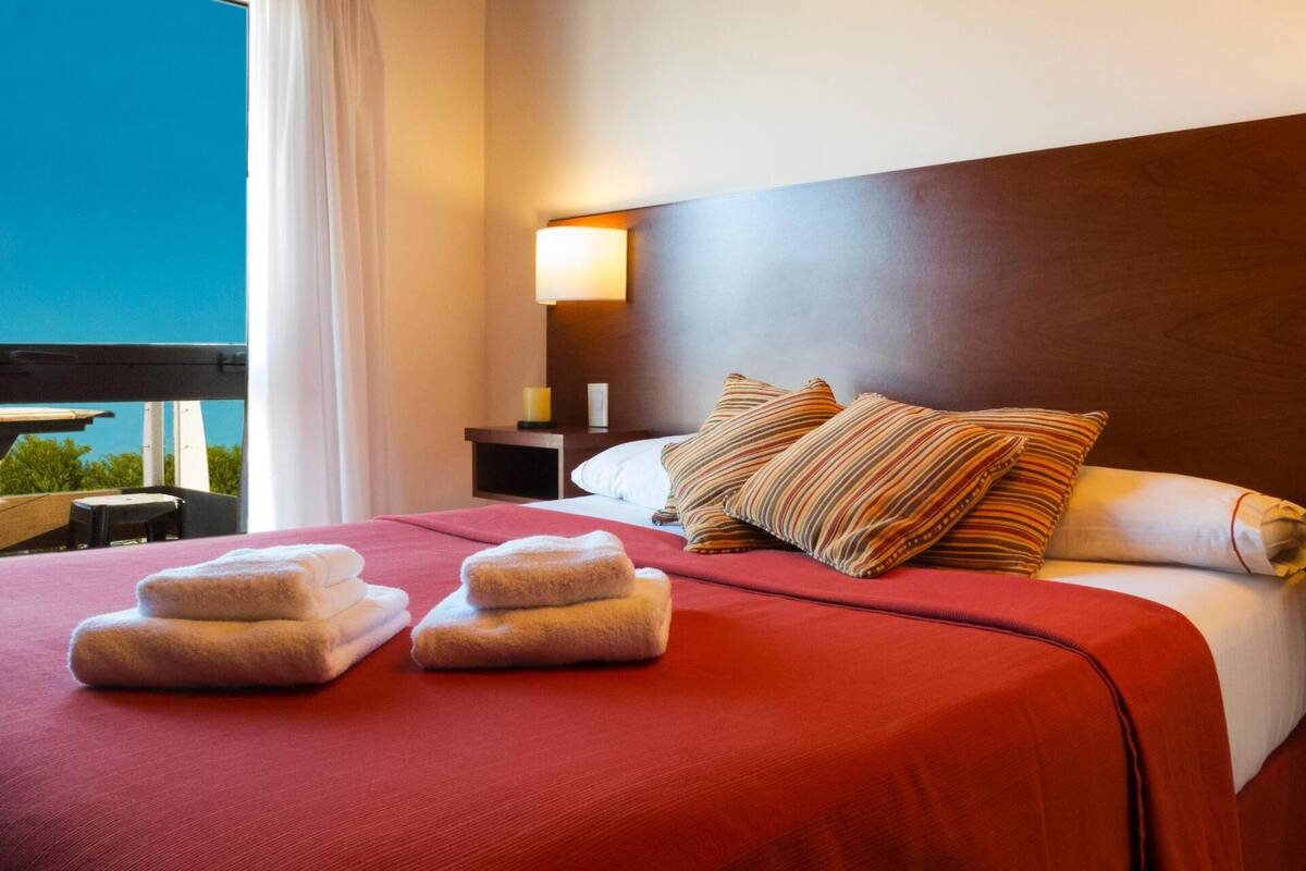 A hotel room with a large bed, red bedding, two pillows, a nightstand with a lamp, and a window with curtains.