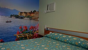 Habitación de hotel con cama estampada con motivos florales, vista a un lago con barcos y un mural de edificios y montañas.