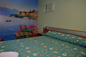 Habitación de hotel con una cama estampada con motivos florales, vista a un lago con barcos y un mural de un pueblo costero.
